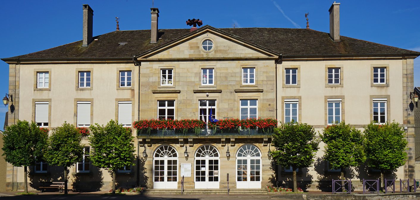 Mairie en pierre avec balcon fleuri et rangée d'arbres taillés en boule sur la place