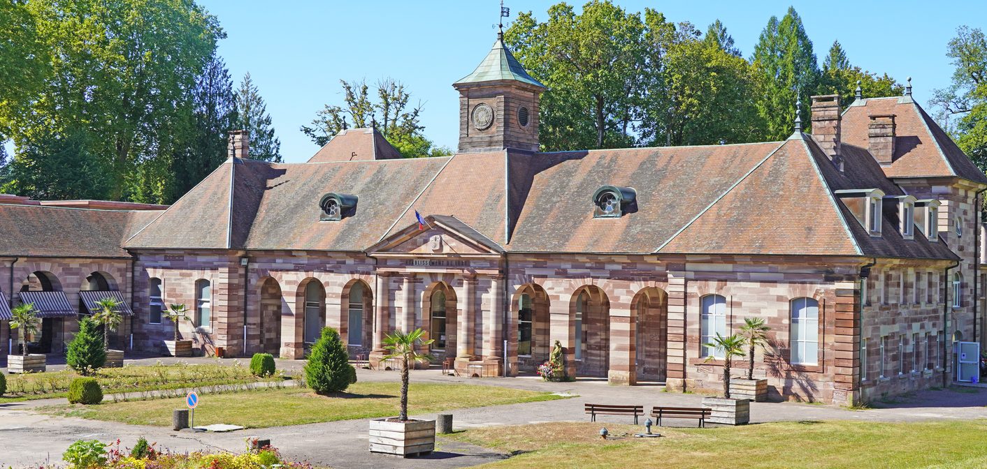 Thermes en pierre rose devant pelouse, topiaires et palmiers en bacs sous un ciel bleu